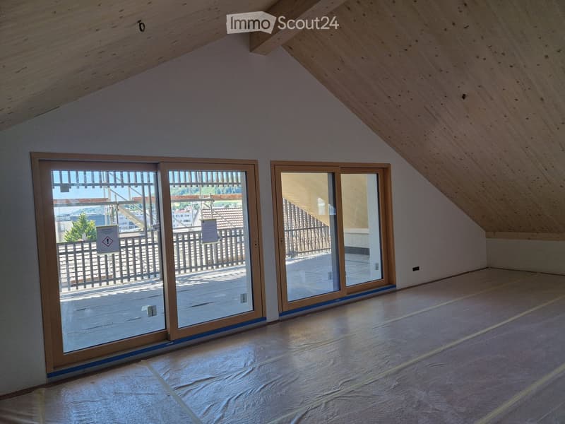Empty attic room with wooden slanted ceilings, large sliding glass doors leading to a balcony, and protective plastic sheeting on the floor.