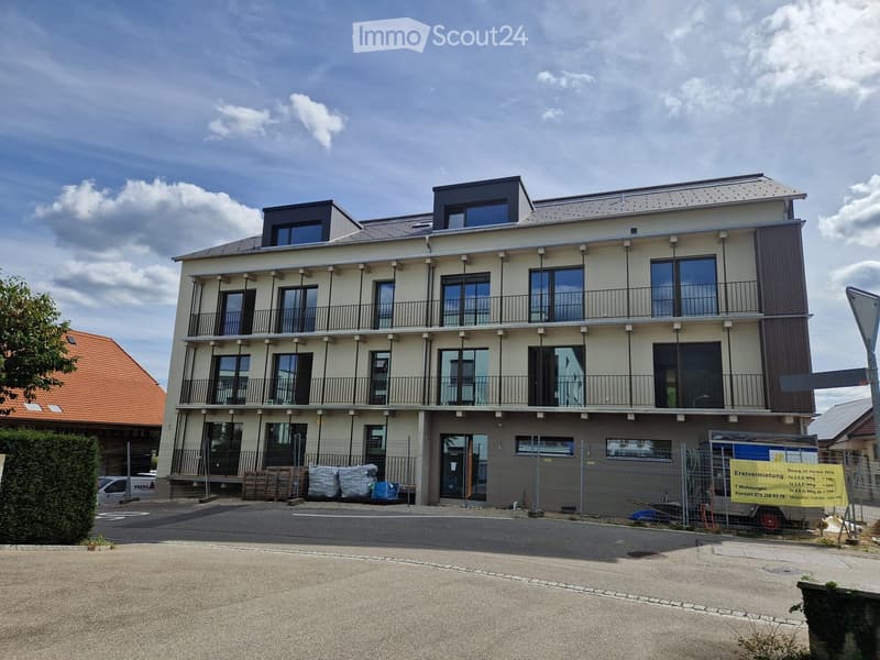 Modern multi-story residential building with balconies, large windows, and a contemporary design. The building has a mix of light-colored walls and dark accents, with a fenced-off area in front.