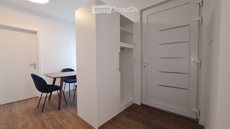 A small, modern room with a wooden floor, a small table, and two blue chairs. The room features white walls, a built-in closet, and a sliding door.