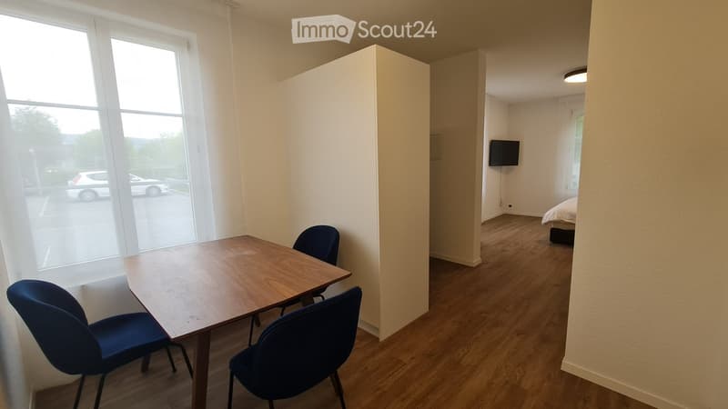 A furnished room with a wooden table, two blue chairs, and a TV screen mounted on the wall. The room has light-colored walls and wooden flooring.
