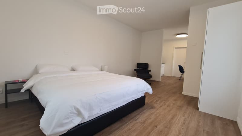 Modern bedroom with a large bed, nightstand, and two chairs. The room features light-colored walls and wooden flooring.
