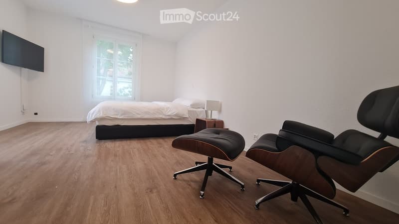 Modern bedroom with wooden flooring, a large bed with white bedding, a nightstand with a lamp, two black leather chairs, and a wall-mounted television.