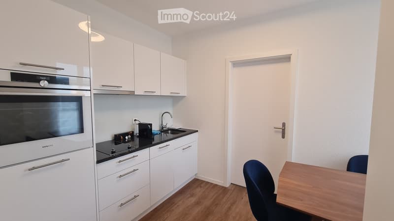 Modern kitchen with white cabinets, stainless steel oven, black countertop, and a small dining area with a table and chair.