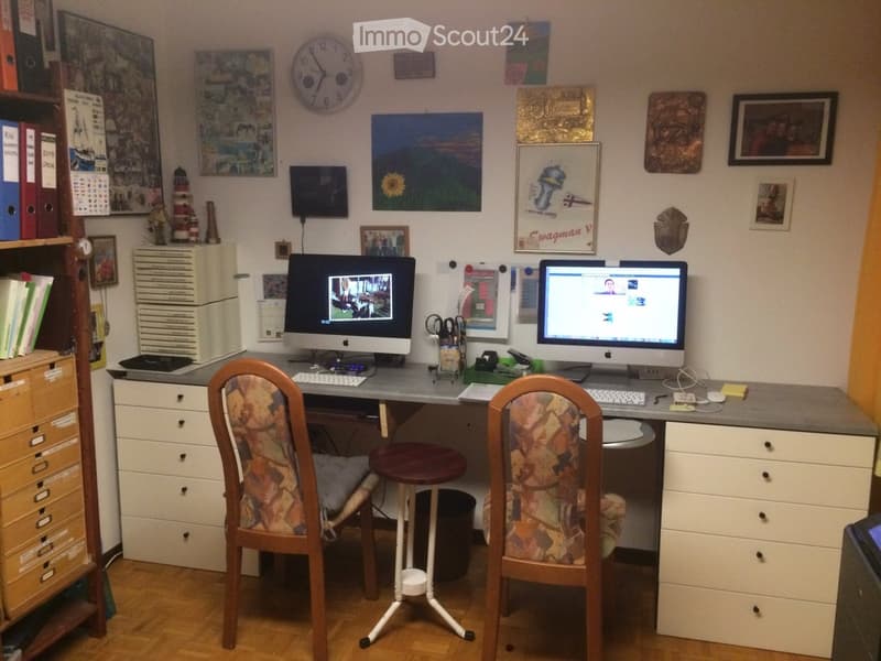 A well-furnished study or office space with a large desk, two chairs, two computer monitors, a filing cabinet, bookshelf, and various wall decorations including paintings and framed pictures.
