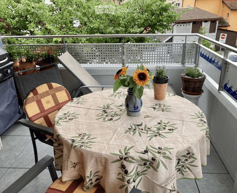 Balcony with a round table covered in a cloth with olive branch design, two chairs, a vase with sunflowers, and several potted plants.