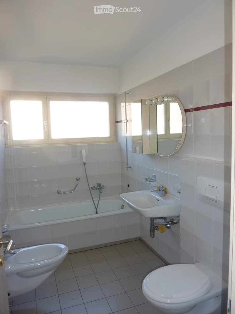 Modern bathroom with white tiles, bathtub, sink, toilet, and a large oval mirror above the sink.