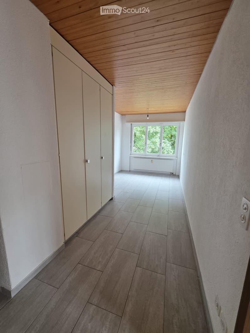 Empty hallway with wooden ceiling, tiled floor, and white walls. A window at the end shows greenery outside.