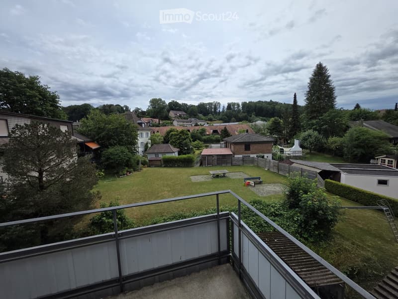 View from a balcony overlooking a well-maintained garden with a lawn, a picnic table, and a bench. The garden is surrounded by a wooden fence and is bordered by neighboring houses and trees.