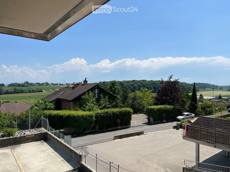 Balcony overlooking a parking area with a car, surrounded by greenery and a house in the background.