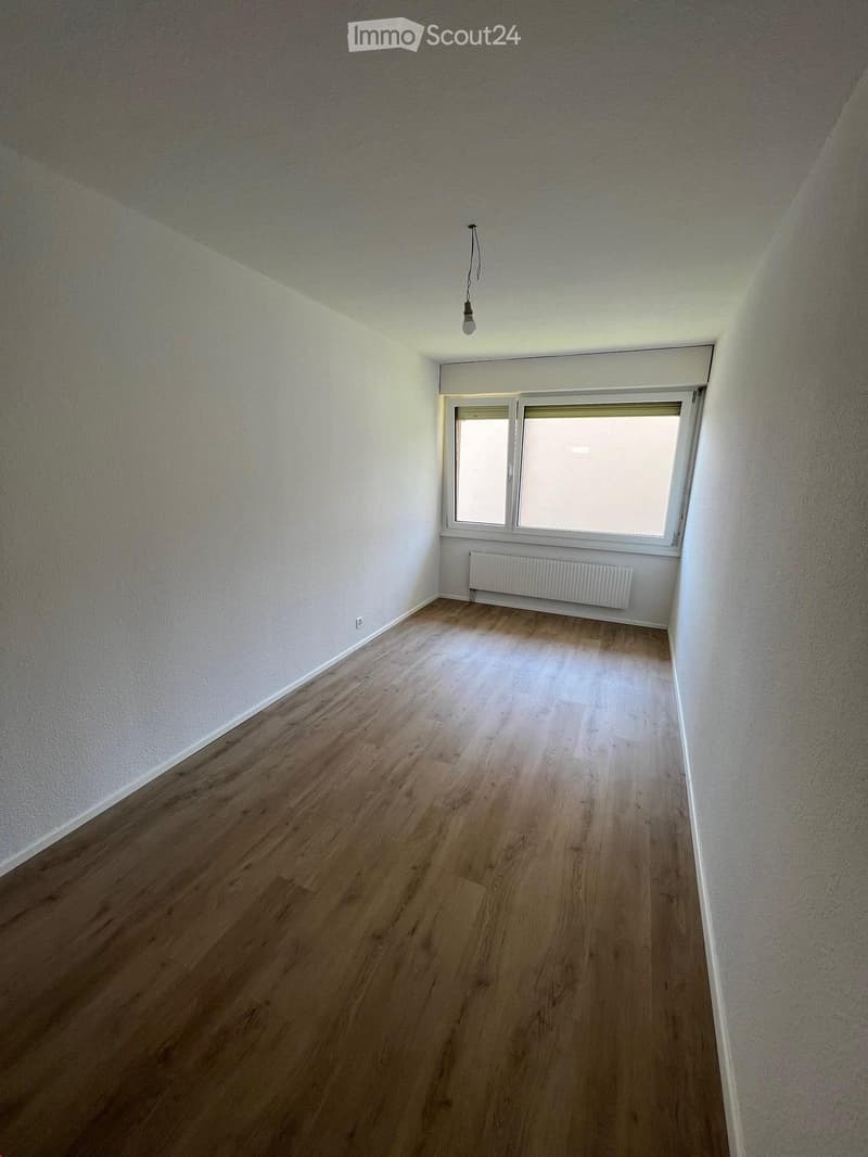 Empty room with wooden flooring, white walls, a window with blinds, a radiator, and a single light fixture hanging from the ceiling.