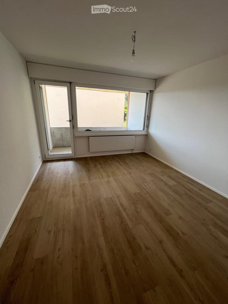 Empty room with wooden flooring, white walls, large window, and sliding door leading outside.