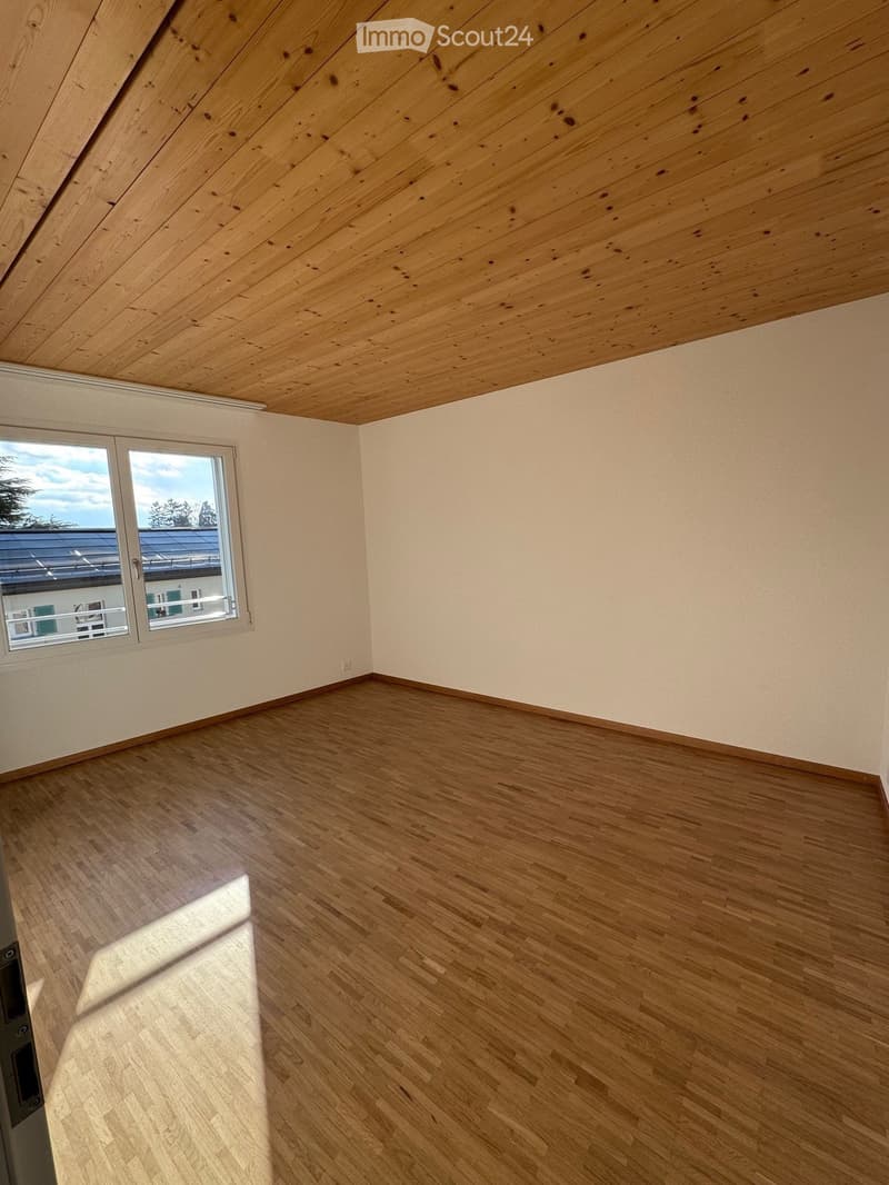 Empty room with wooden flooring and a slanted wooden ceiling. Large window with a view of the outside.