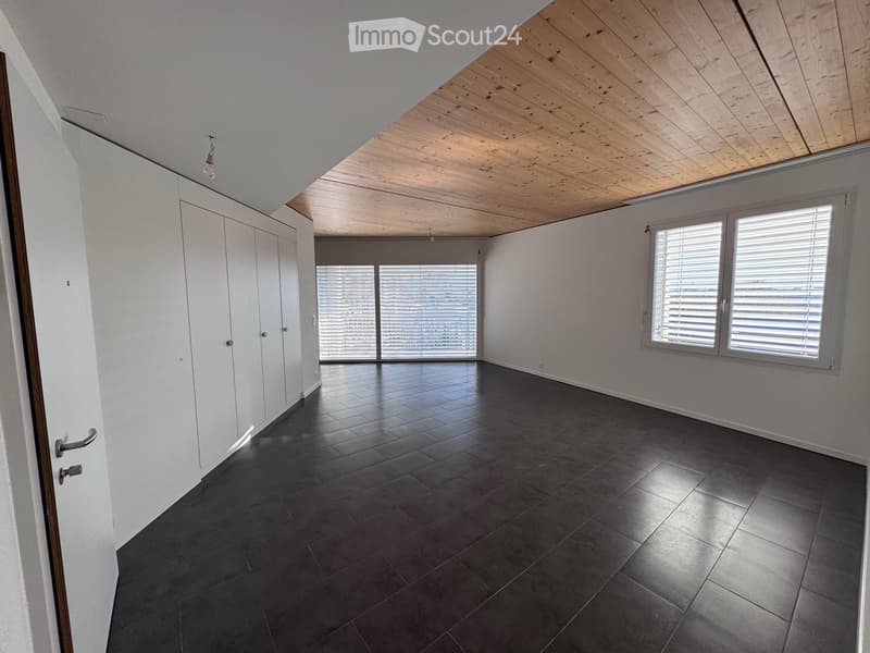 Empty room with dark tiled floor, white walls, large windows with blinds, and a wooden slanted ceiling.
