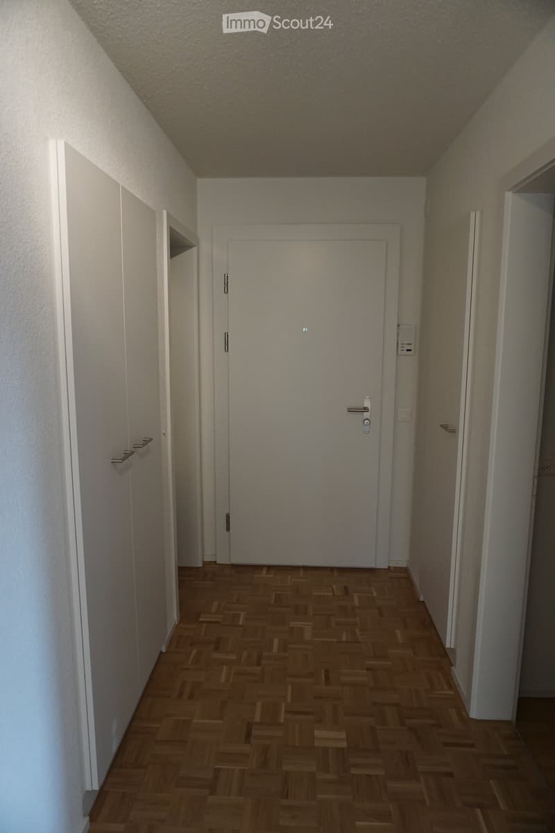 A narrow hallway with wooden flooring and white walls, featuring a central door and several wall-mounted shelves on the left side.