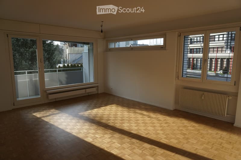 Empty room with wooden flooring, large windows, radiators, and a sliding glass door leading to a balcony.