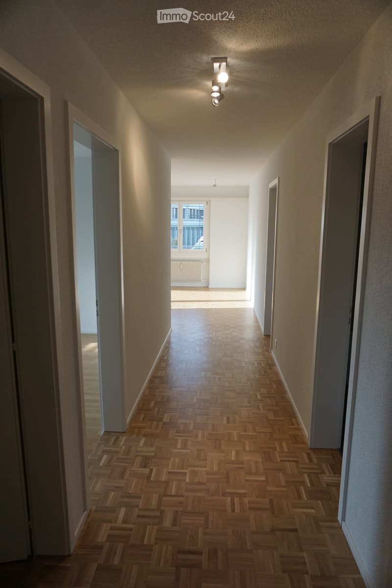 Empty hallway with wooden herringbone flooring, beige walls, and recessed lighting. Doors lead to adjacent rooms.