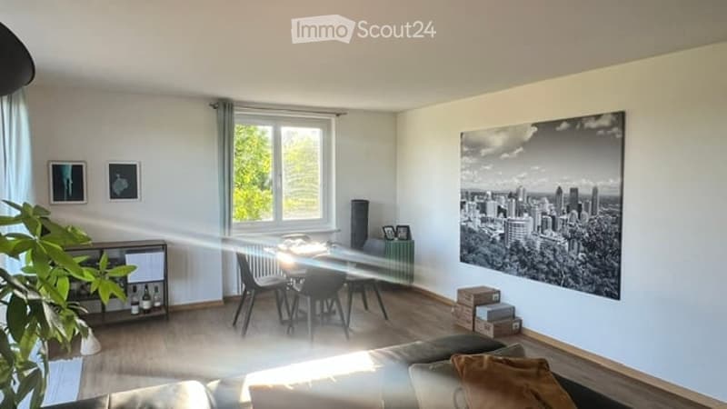 Bright living room with wooden flooring, large window, sofa, coffee table, dining area with table and chairs, and artwork on the wall.
