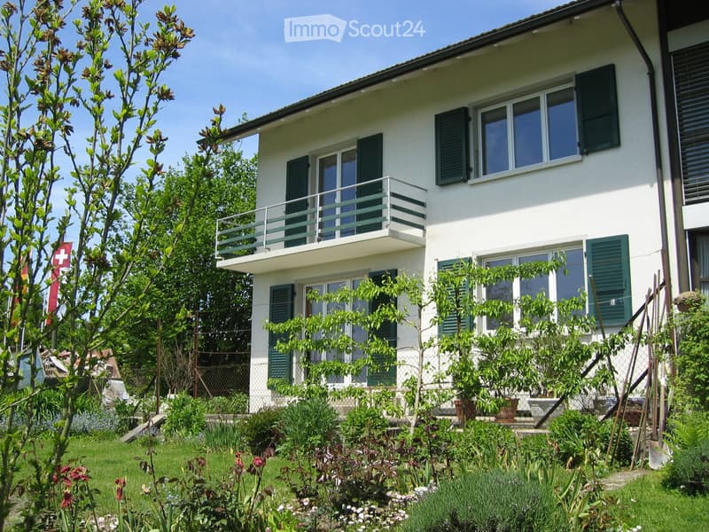 Two-story white house with green shutters, large windows, and a balcony on the second floor. The house is surrounded by a well-maintained garden with various plants, trees, and a Swiss flag.