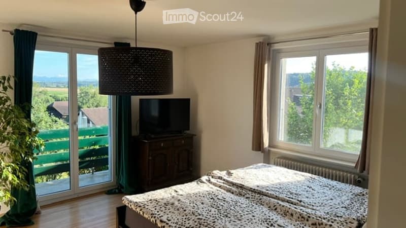Bright bedroom with large windows offering a scenic view, featuring a bed with a patterned duvet, a television on a wooden dresser, and a floor lamp.