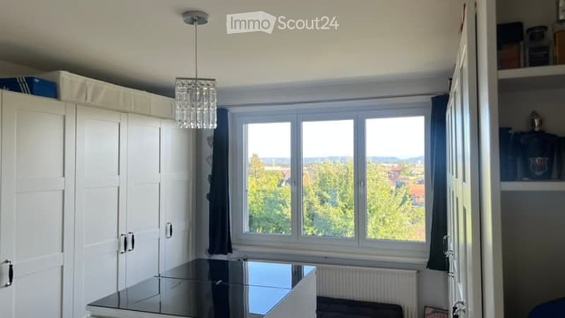 A well-lit room with a large window offering a scenic view, featuring a white wardrobe, a sleek black table, a chair, and a curtain partially drawn.