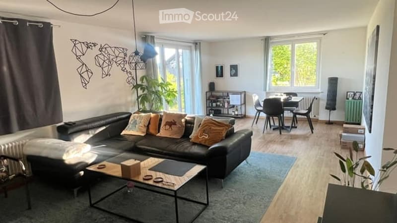 Modern living and dining area with a black leather sofa, coffee table, dining table with chairs, and decorative plants. The space features large windows, a world map wall decoration, and a sleek lamp.