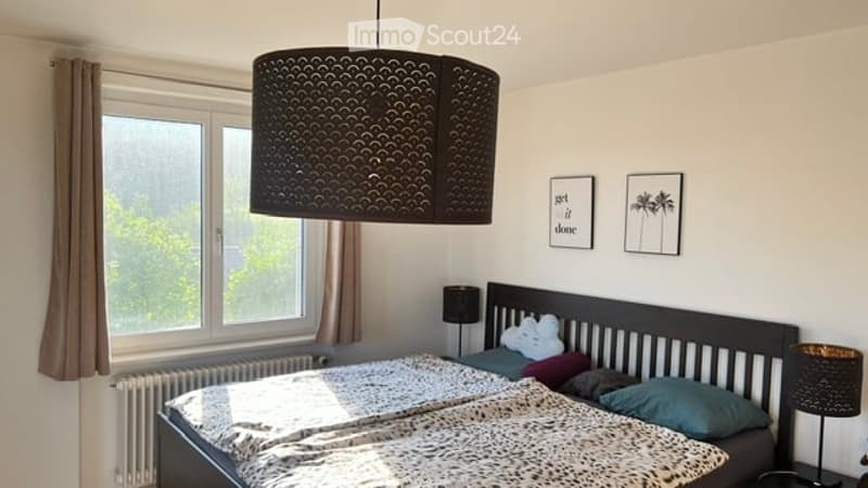 Cozy bedroom with a large window, a dark wooden bed with a patterned blanket, pillows, a nightstand with a lamp, and two picture frames on the wall.