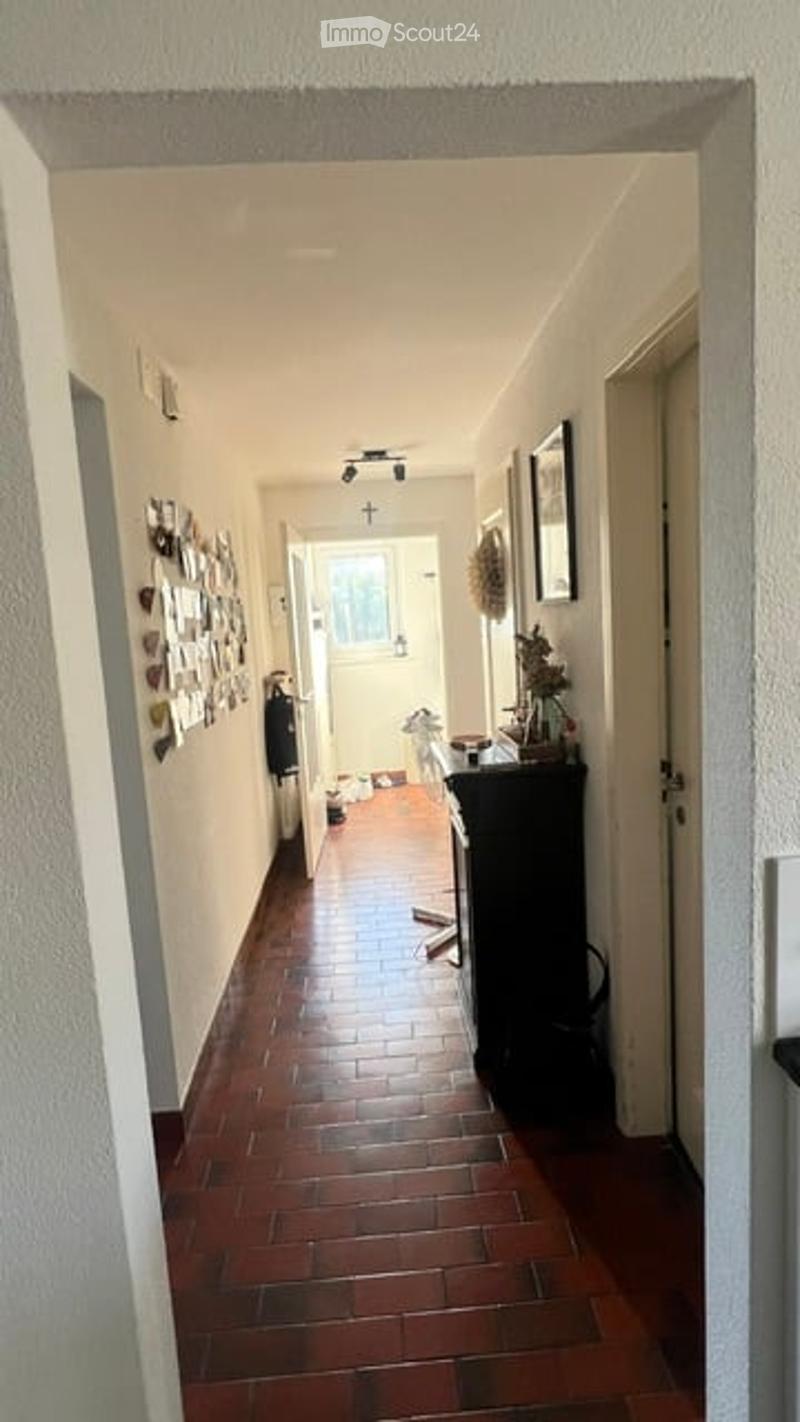 A narrow hallway with red brick flooring, white walls, and several framed pictures and decorations on the left side. A small table with items is visible on the right side.