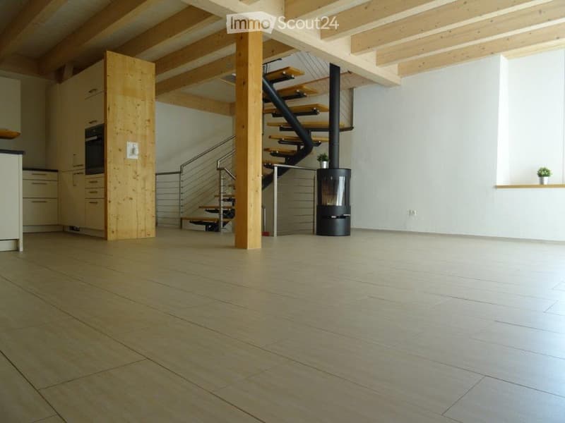 Open-plan space with exposed wooden beams, a staircase leading to an upper level, and a modern wood-burning stove. The floor is light-colored tile, and the walls are white.