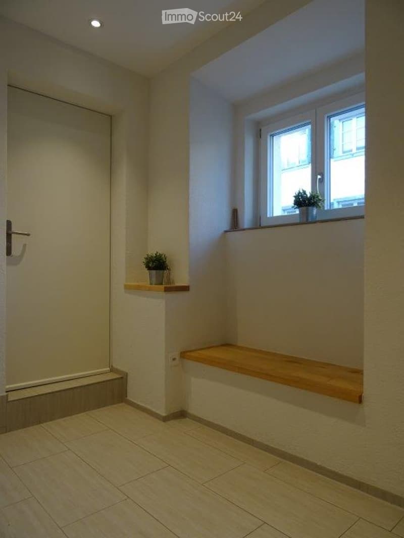Empty hallway with tiled floor, white walls, a small wooden bench, and a small shelf with a potted plant near a window.