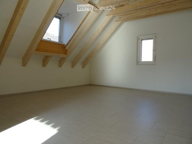 Empty attic room with wooden beams, wooden floor, and two windows allowing natural light.