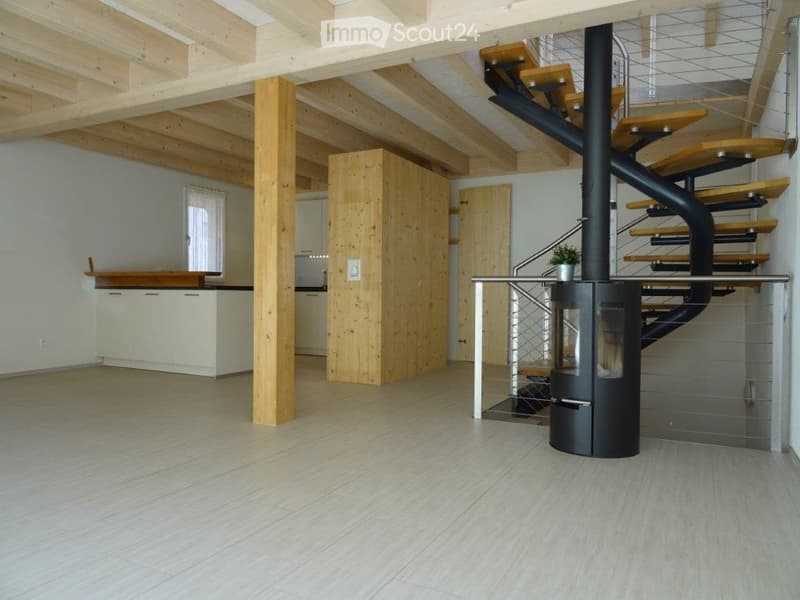Open-plan living and kitchen area with exposed wooden beams, a modern black fireplace, a spiral staircase, and white kitchen cabinets.