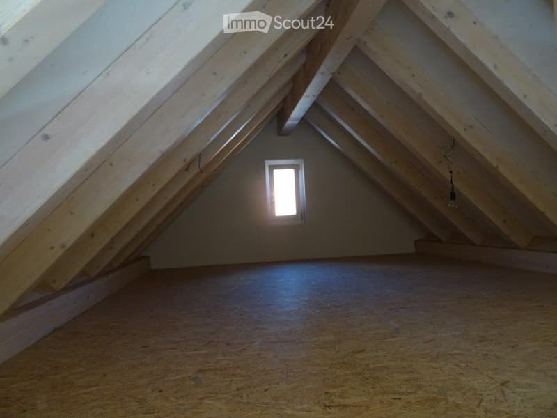 Empty attic space with wooden beams, exposed wooden floor, and a small window letting in natural light.
