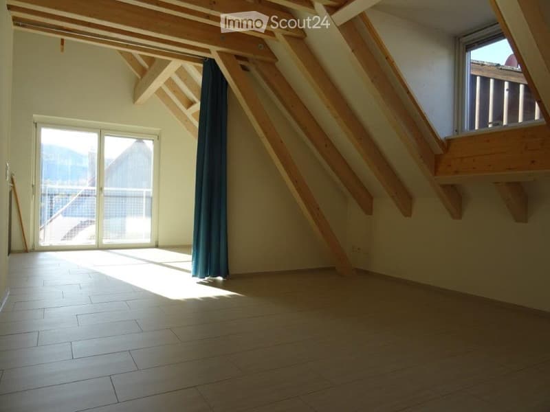 Bright, empty attic room with wooden beams, large windows, and a sliding glass door leading to a balcony with a scenic view.