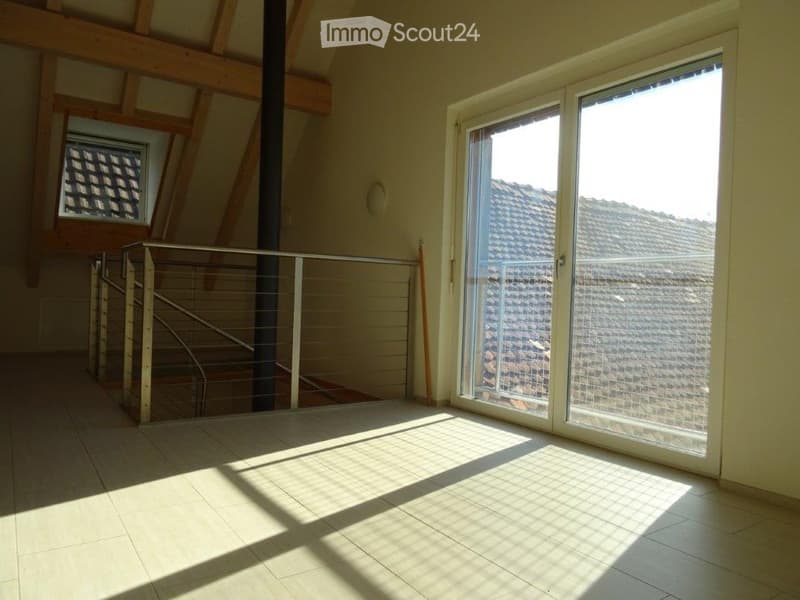Bright, empty room with large windows, wooden beams, and a staircase leading upwards. The floor is tiled, and the walls are painted a light color.