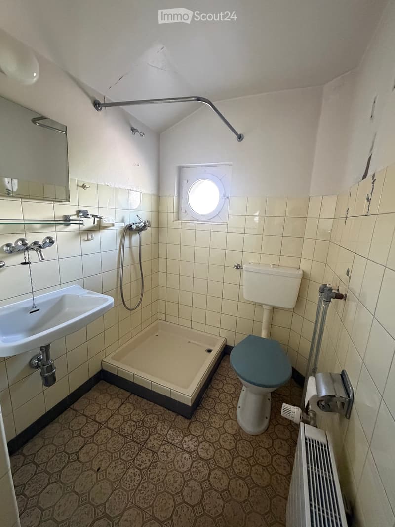 Small bathroom with a sink, toilet, shower, mirror, and radiator. The walls are tiled, and the floor has a hexagonal pattern.