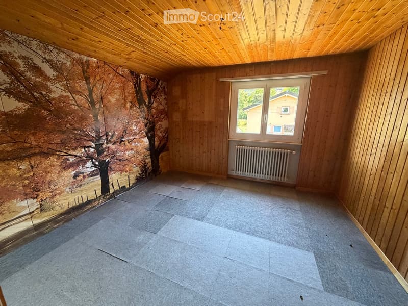 Empty room with wooden walls and ceiling, large window, radiator, and mural of a landscape on one wall.