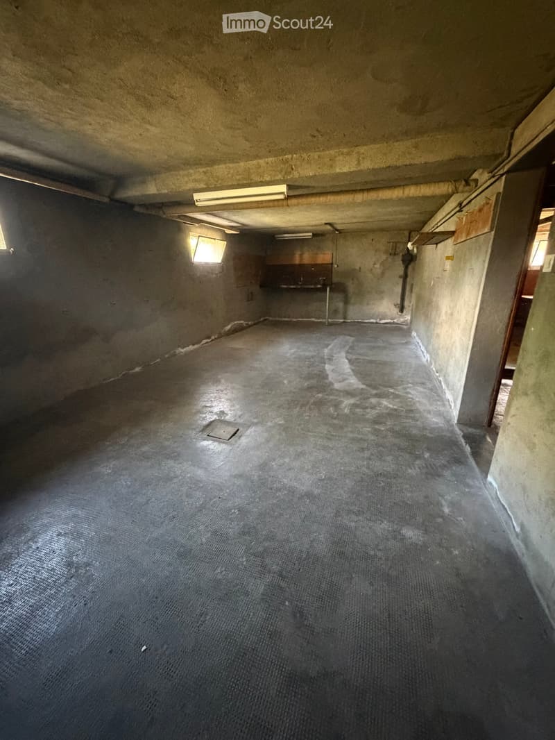 Empty basement space with concrete walls, floor, and ceiling. Features include exposed pipes, a small window, and a metal grate on the floor.