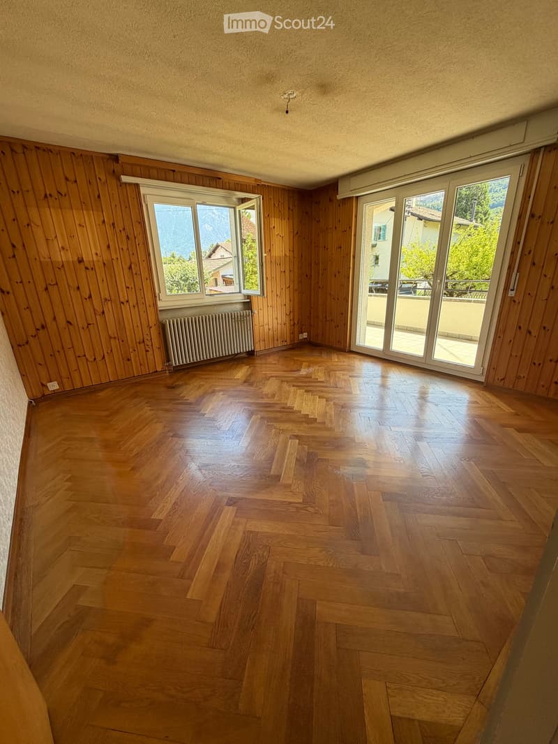 Empty room with wooden flooring, wooden panel walls, large windows, and a radiator. No furniture or equipment present.