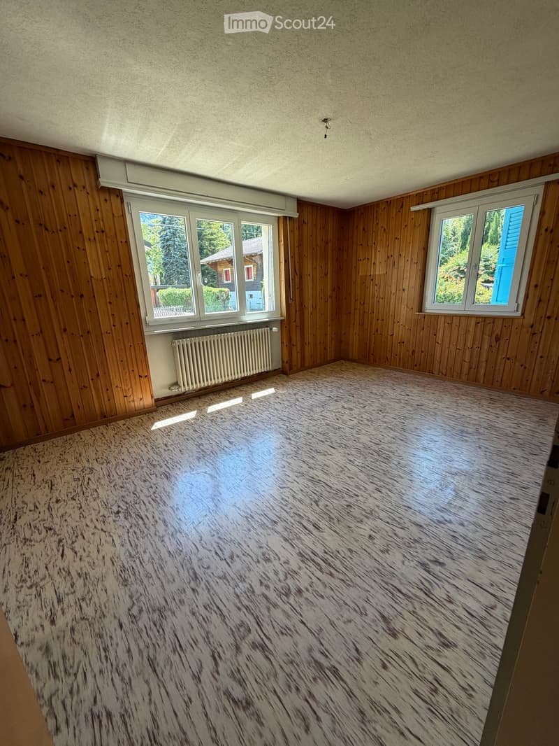 Empty room with wooden walls, laminate flooring, and large windows offering a view of the outside. A radiator is visible under one window.