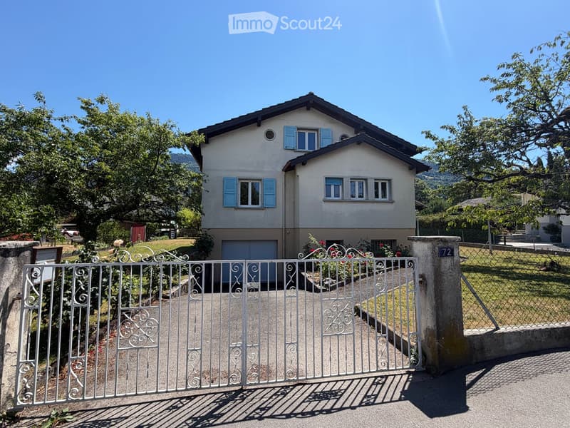 Two-story house with light-colored walls, blue shutters, and a garage. The property is surrounded by a white metal gate, greenery, and trees.
