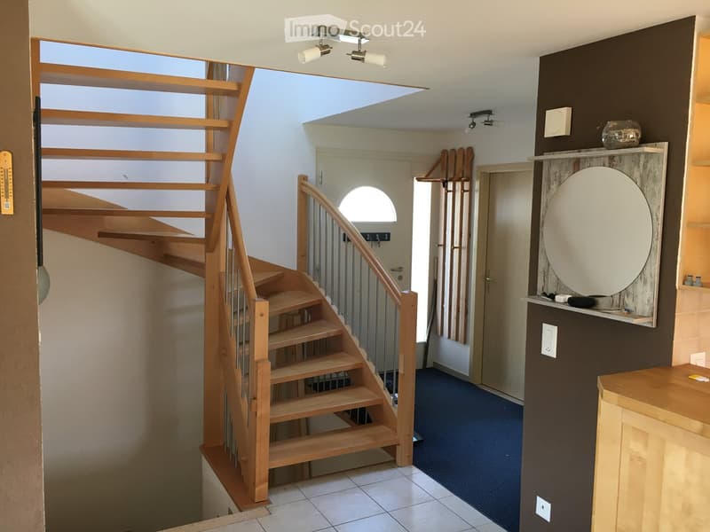 Modern hallway with wooden staircase, metal railing, and built-in shelf with decorative items. The space features tiled flooring, recessed lighting, and a door leading to another room.