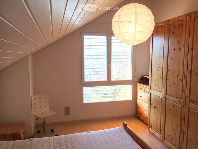 Bright bedroom with wooden floor, slanted ceiling, large windows with blinds, bed, chair, wardrobe, and pendant light.