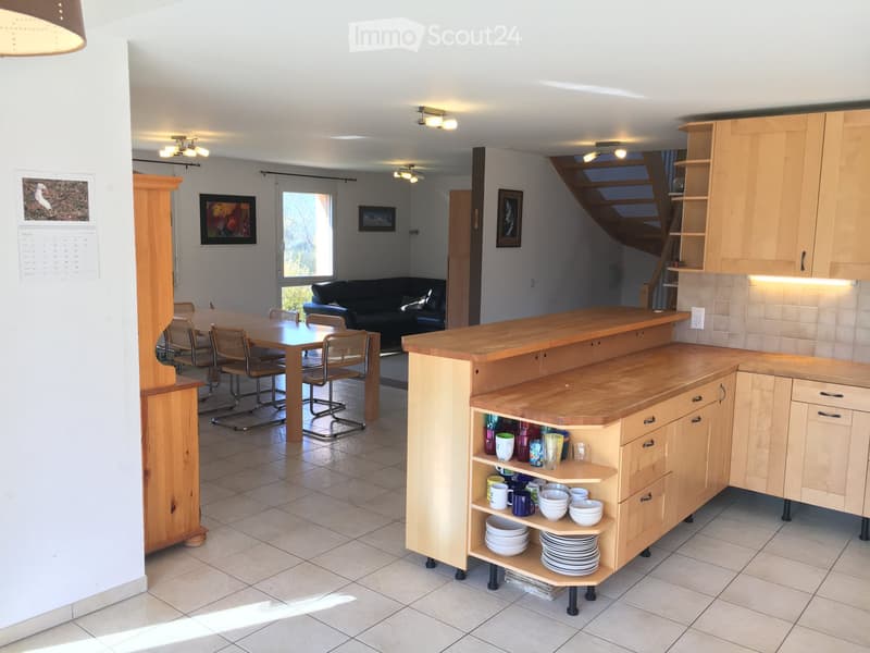 Open-plan living and dining area with a kitchen island, dining table, chairs, and a sofa. The kitchen features wooden cabinets and a countertop with dishes and cups.
