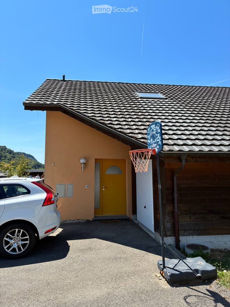 Single-story house with a tiled roof, yellow front door, and a basketball hoop in the driveway. A white car is parked nearby.