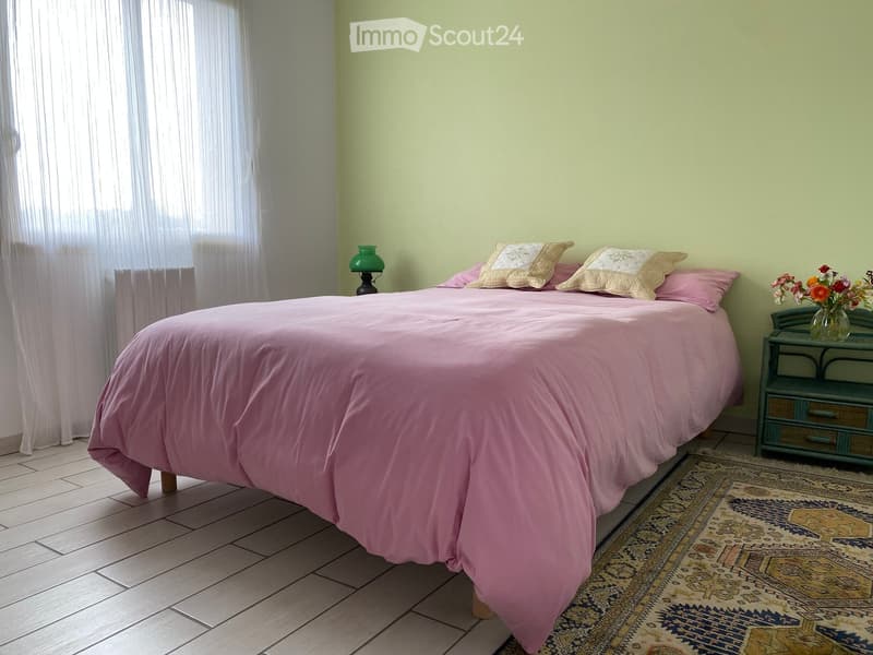 Cozy bedroom with a double bed covered in pink bedding, two decorative pillows, a nightstand with a table lamp, a vase with flowers, and a patterned rug on the floor.