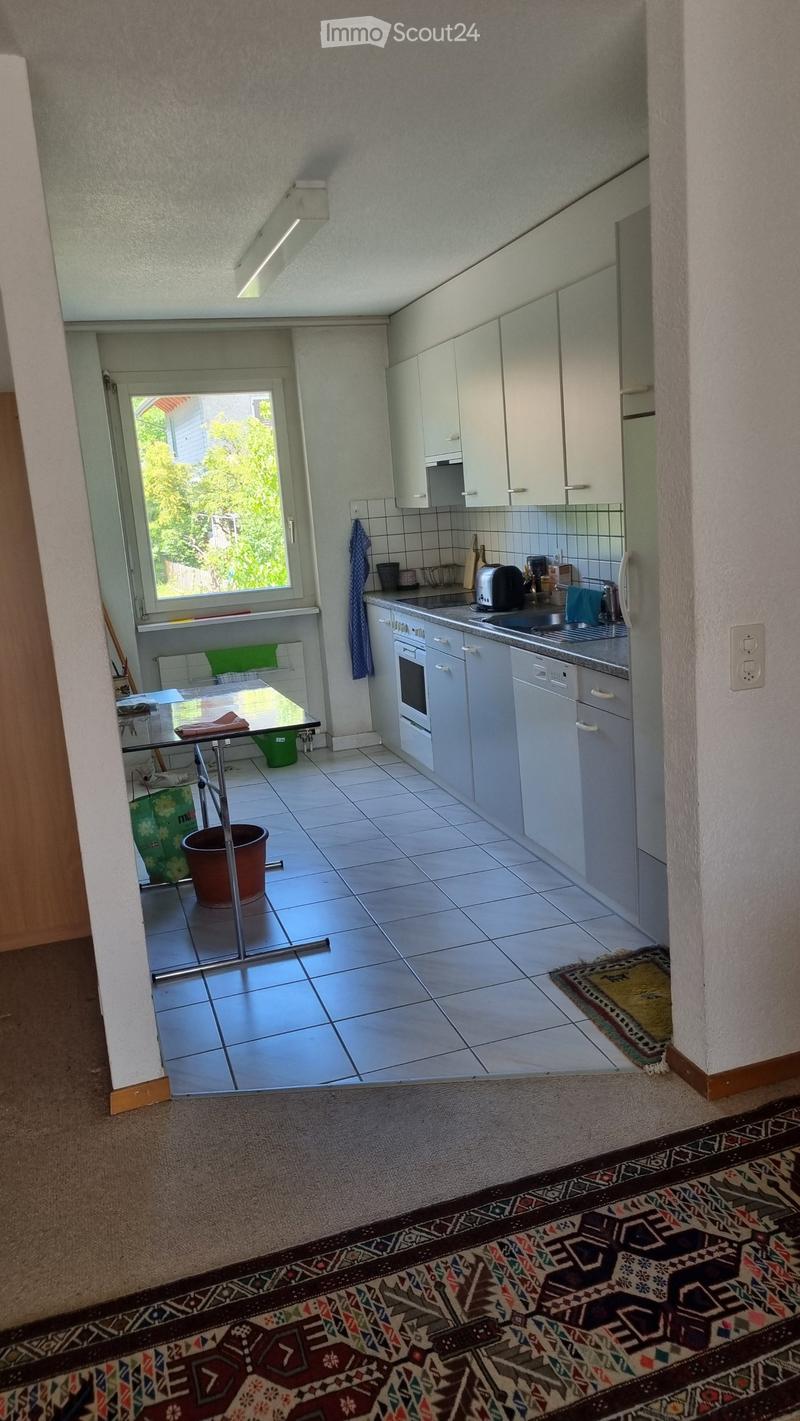 The kitchen features white cabinets, a tiled floor, and a window with a view of greenery. A sink, oven, and dishwasher are visible, along with a small table and various kitchen items.