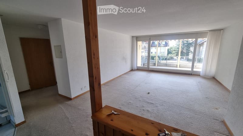 Empty room with carpeted floor, large window with white curtains, wooden door, and a small wooden table with keys and a phone on it.