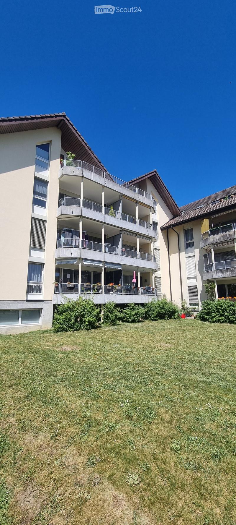 Multi-story residential building with multiple balconies, surrounded by green lawn and bushes.