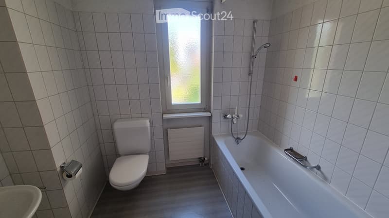 Bathroom with tiled walls and floor, featuring a toilet, bathtub, and a window with frosted glass. Includes a radiator and a toilet paper holder.