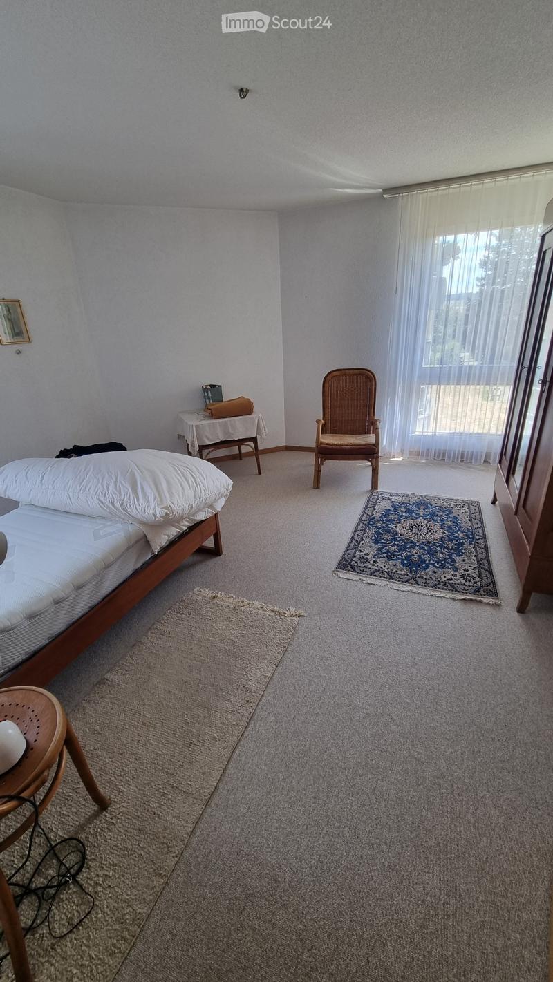 A well-lit bedroom with a bed, a wooden chair, a small table with a cloth, and two rugs on the carpeted floor. The room features large windows with sheer curtains and a framed picture on the wall.